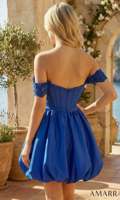 Wearing the Amarra 89705 short homecoming dress, a woman with long blonde hair stands outdoors near stone columns and potted plants. The royal blue, off-the-shoulder fit-and-flare dress features lace sleeves and a stylish corset bodice.