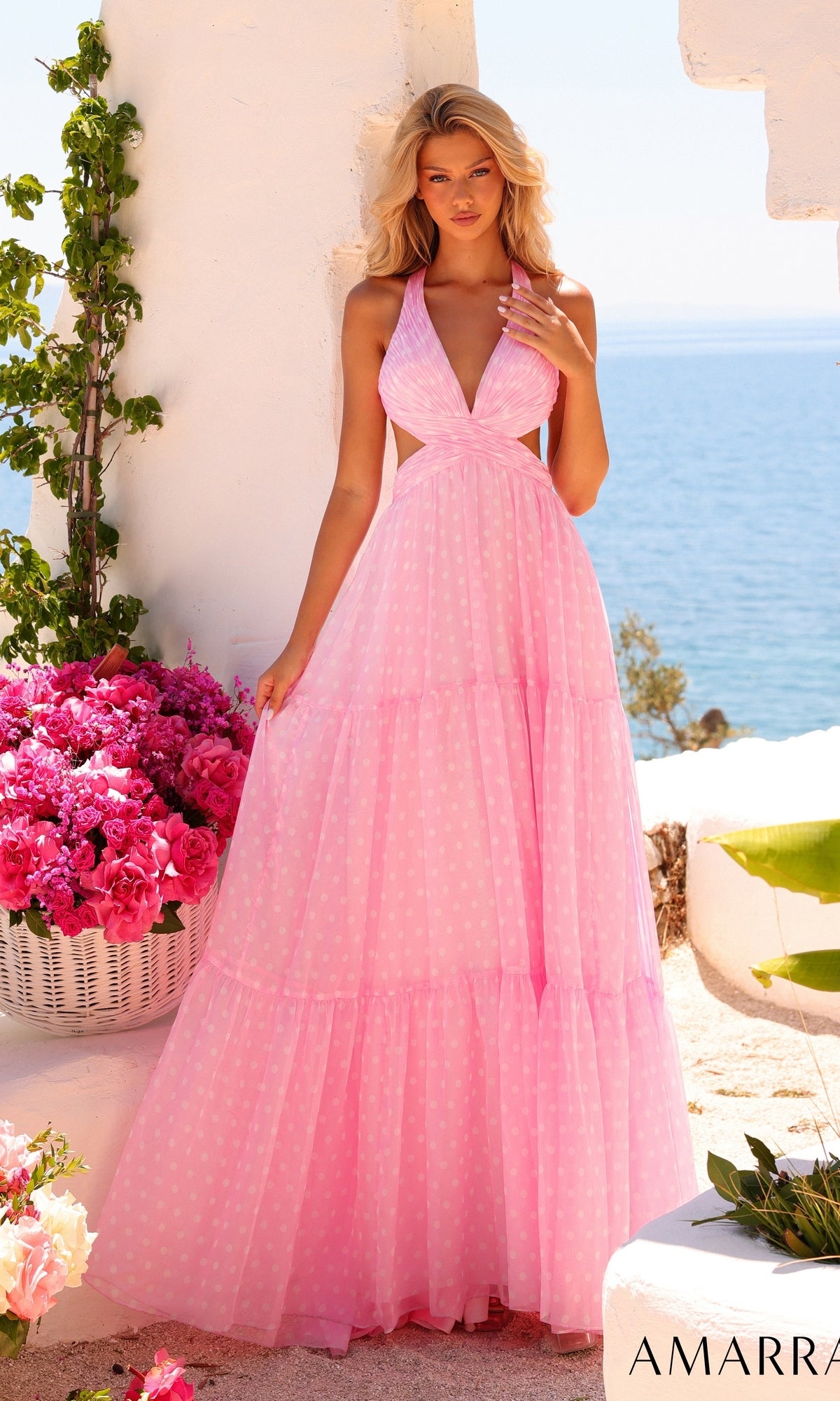 A woman wearing the Amarra 89730 long prom dress in light pink, featuring a halter neckline and charming polka dot print, stands outdoors near white walls with pink flowers and the sea in the background.