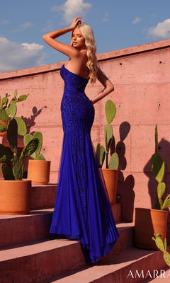 Wearing the Amarra 89736 long prom dress, a woman poses on outdoor steps beside potted cacti, her hand resting on her head as she stands against a pink concrete wall and blue sky. The strapless sweetheart gown shimmers with royal blue sequins.
