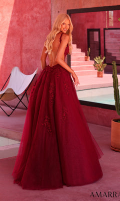 A woman stands outdoors near stairs, her long blonde hair cascading down the low-cut back of the Amarra 89786 Long Prom Dress—a deep red, floor-length gown adorned with delicate floral lace appliqués. Modern furniture and potted plants are visible in the background.