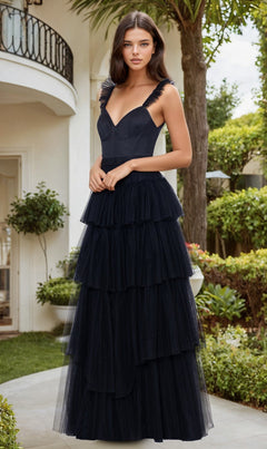 A woman stands outdoors in a garden, wearing the Blondie Nites 4819BN Ruffled Tiered Long Prom Dress—sleeveless, floor-length, and black—with greenery and a white building in the background.