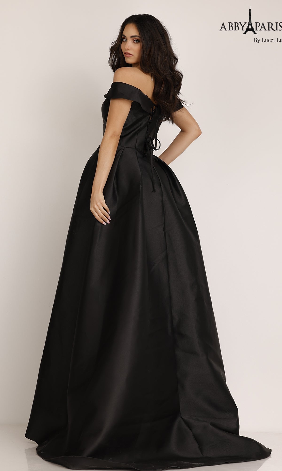 A woman in the Classic Mikado Prom Ball Gown, Abby Paris 90023—a black, off-the-shoulder, floor-length dress—poses against a plain background with one hand on her hip.