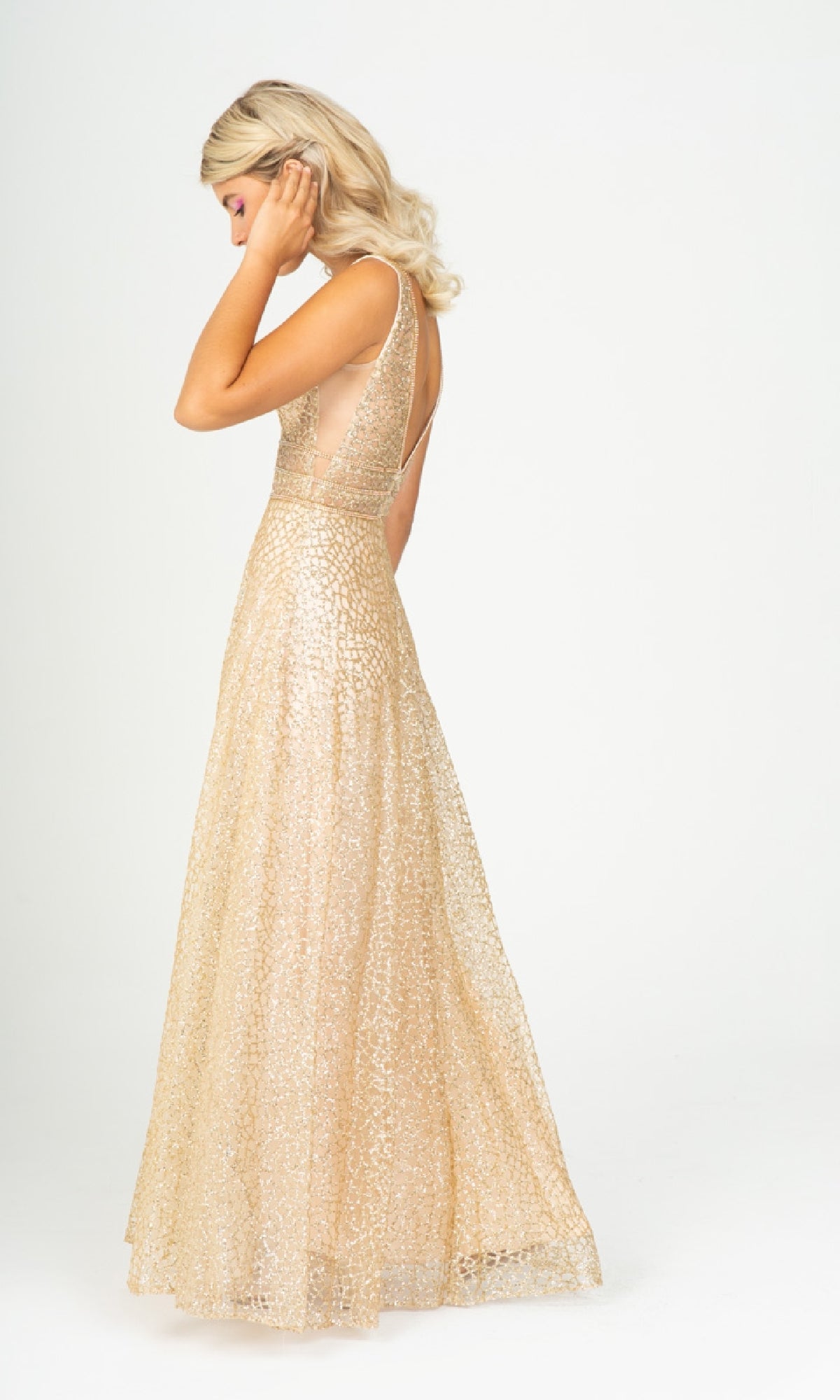 A woman in the Sleeveless Long Glitter Formal Dress: Eureka 9005, a floor-length gold patterned gown, stands against a plain white background, facing slightly away and touching her hair.
