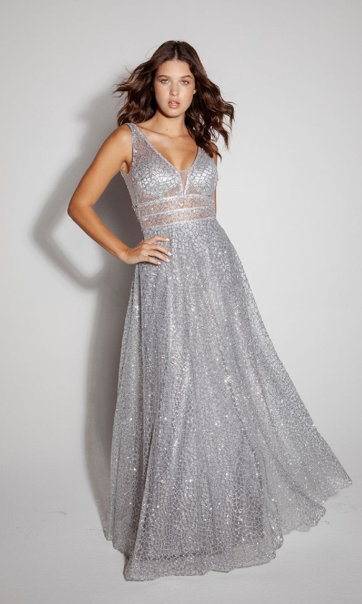 A woman poses against a plain backdrop wearing the Eureka 9005, a sleeveless long glitter formal dress with silver sequins, a deep V-neck, and sheer waist panel details.