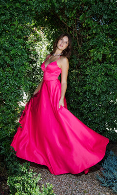 A woman poses outdoors in the Strappy-Back Long Satin Prom Dress: Eureka 9008, her vibrant pink gown flowing gracefully as she stands amid lush green foliage.