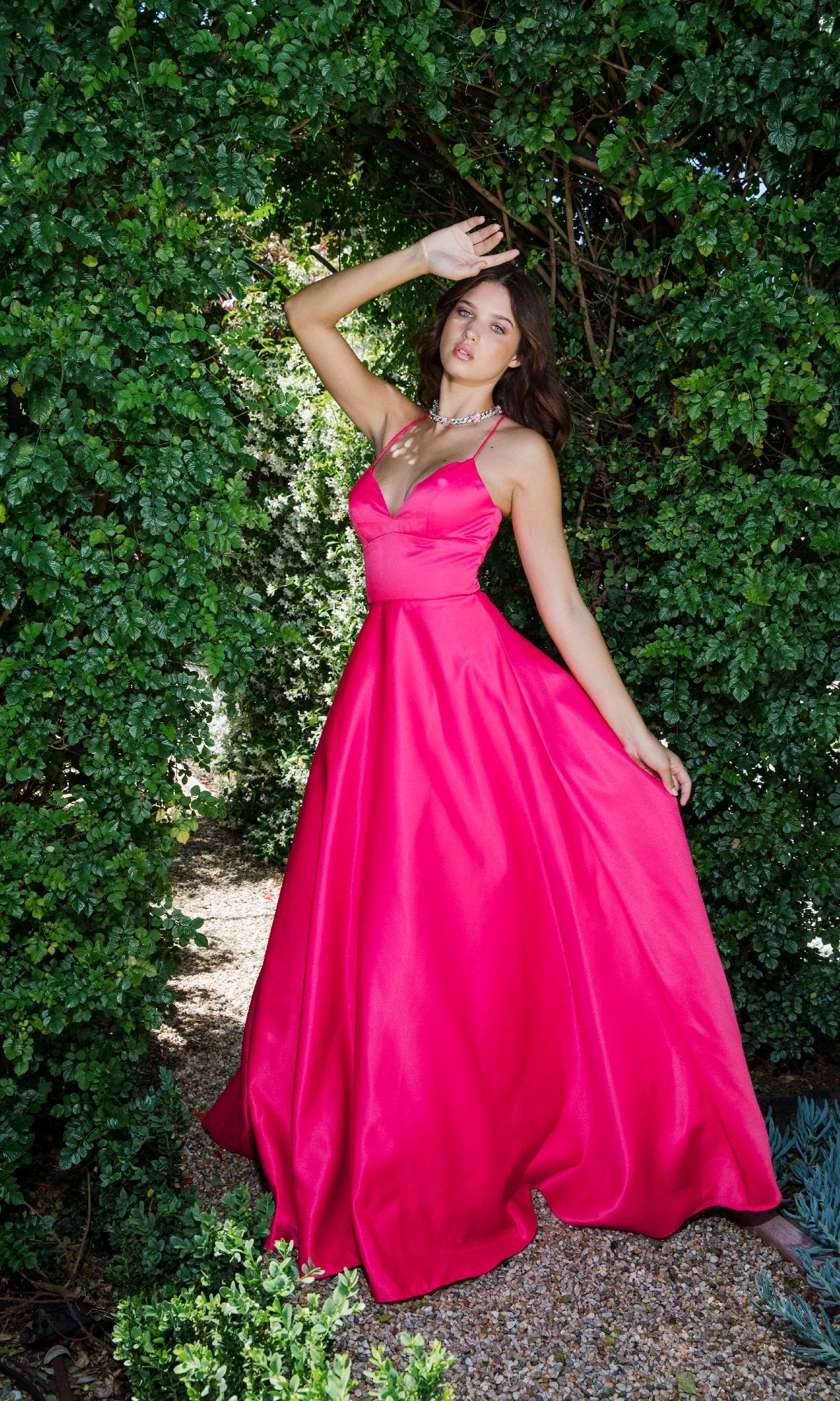 A woman stands outdoors between green bushes, wearing the Strappy-Back Long Satin Prom Dress: Eureka 9008 in bright pink, striking a pose with one arm raised as sunlight filters through the leaves.