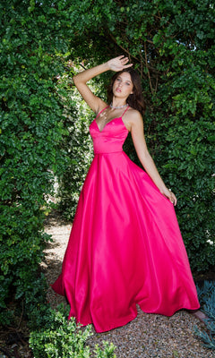 A woman stands outdoors between green bushes, wearing the Strappy-Back Long Satin Prom Dress: Eureka 9008 in bright pink, striking a pose with one arm raised as sunlight filters through the leaves.