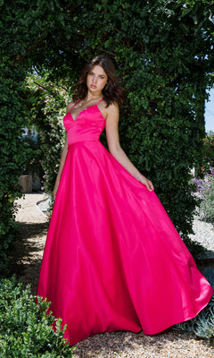 A woman wearing the Strappy-Back Long Satin Prom Dress: Eureka 9008 in bright pink stands outdoors under a leafy archway, holding out the skirt of her floor-length gown.