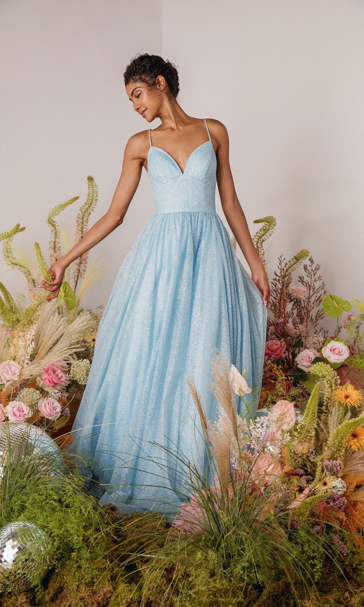 A woman in the Strappy-Back Glitter Prom Ball Gown: Eureka 9009 stands among flowers and greenery, looking down as she gently holds her sparkling light blue dress.