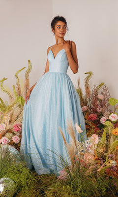 A woman in the Strappy-Back Glitter Prom Ball Gown: Eureka 9009, a light blue sparkly evening dress with thin straps, stands among flowers and grasses in a softly lit room.