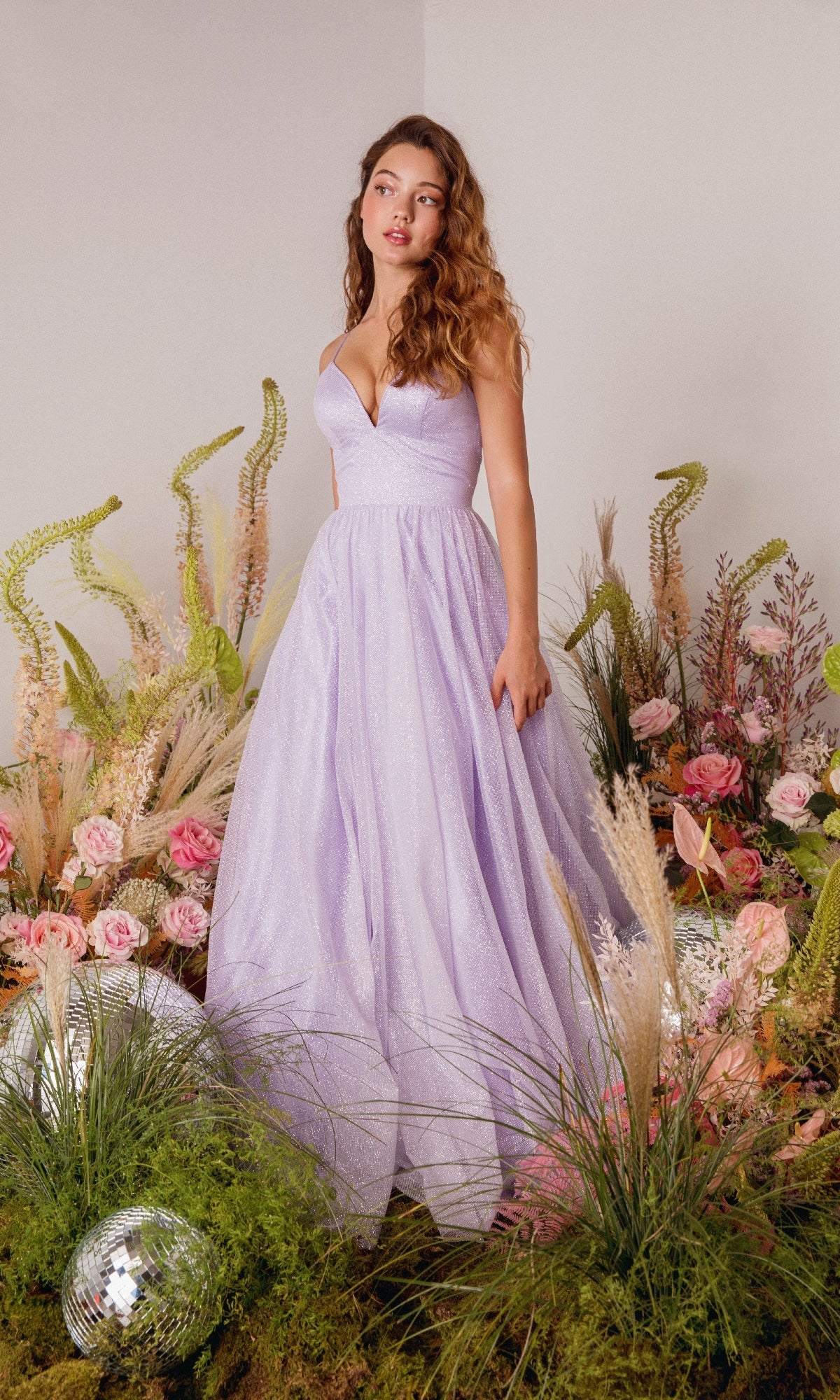 A woman wearing the Strappy-Back Glitter Prom Ball Gown: Eureka 9009 stands among green plants, grasses, and pink flowers, with disco balls on the ground nearby.