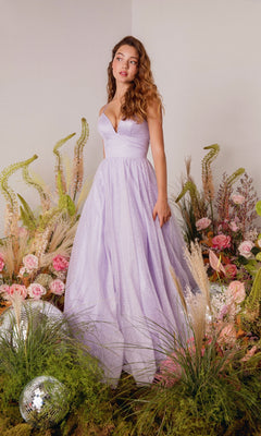 A woman wearing the Strappy-Back Glitter Prom Ball Gown: Eureka 9009 stands among green plants, grasses, and pink flowers, with disco balls on the ground nearby.