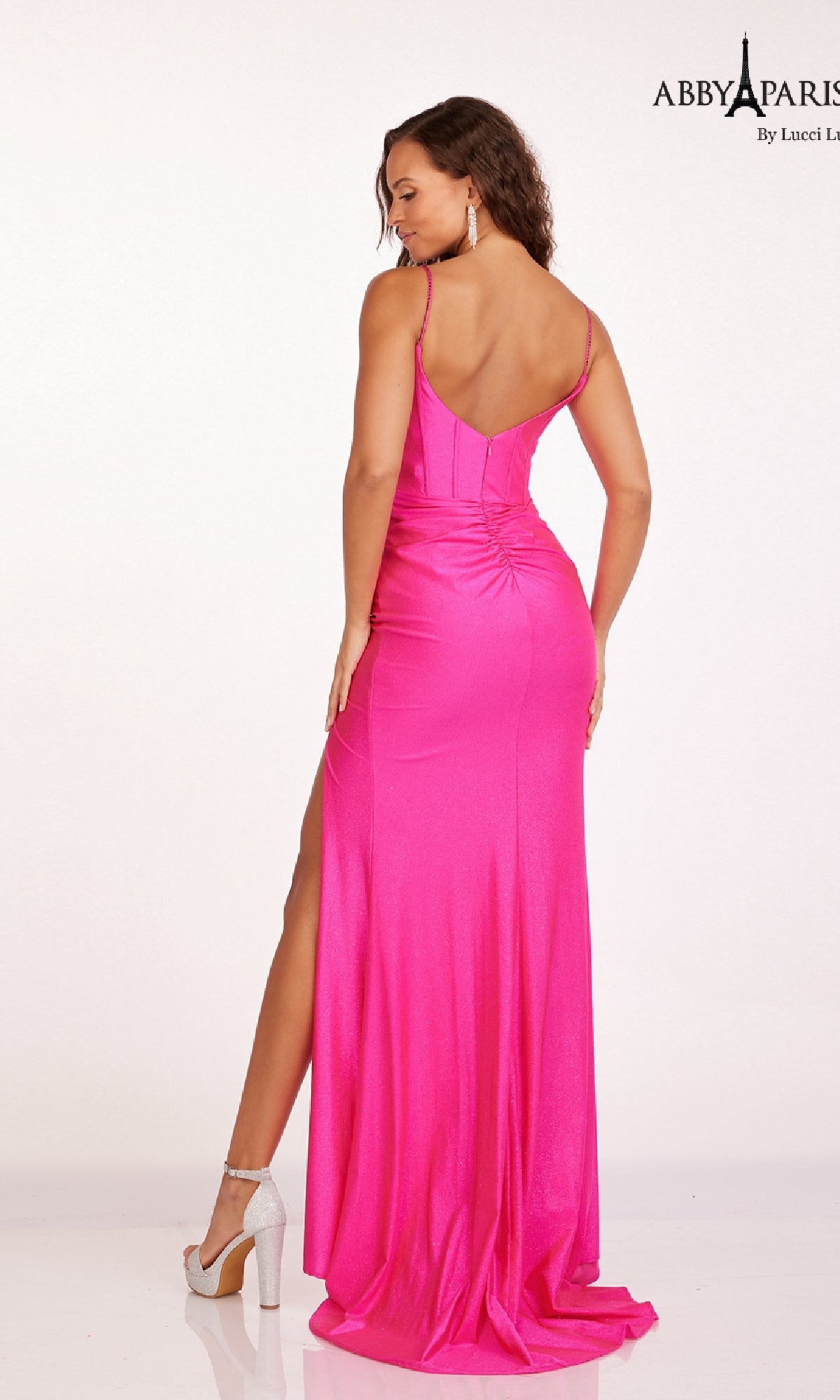 A woman wearing the Abby Paris 90203 Ruched Corset Glitter Prom Dress in bright pink, featuring thin straps, a floor-length silhouette, side slit, and elegant ruching detail, stands with her back to the camera.