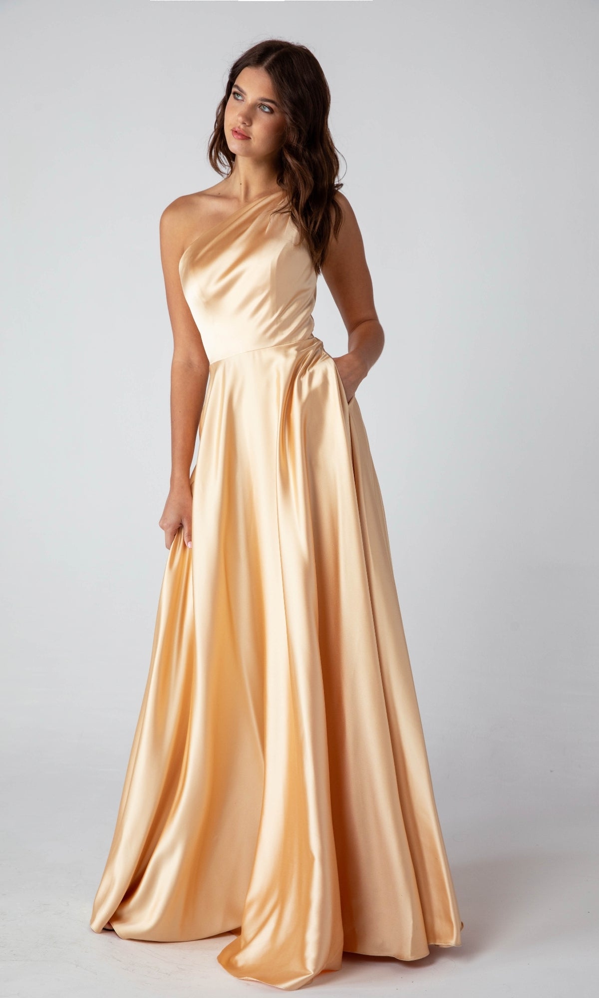 A woman poses in a studio wearing the Eureka 9025, a floor-length, one-shoulder satin formal dress in light peach—an elegant choice for evening events.