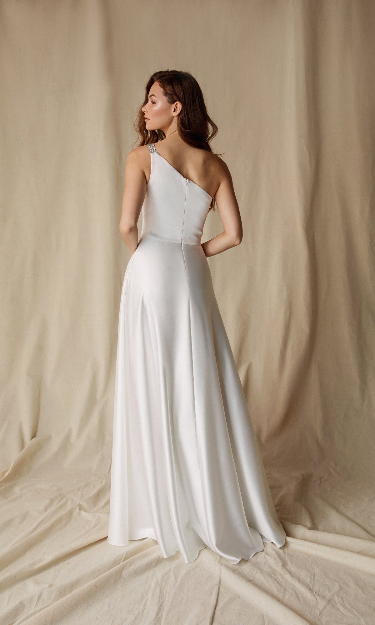 A woman faces away from the camera in front of a beige fabric backdrop, wearing the Eureka 9025 One-Shoulder Long Satin Formal Dress in white.