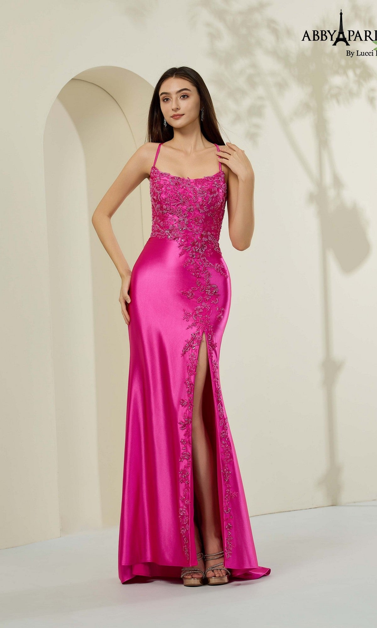 A woman in the Abby Paris 90357 long prom dress in fuchsia, featuring floral embroidery, a beaded bodice, and a thigh-high slit, stands in a minimal light-colored setting.