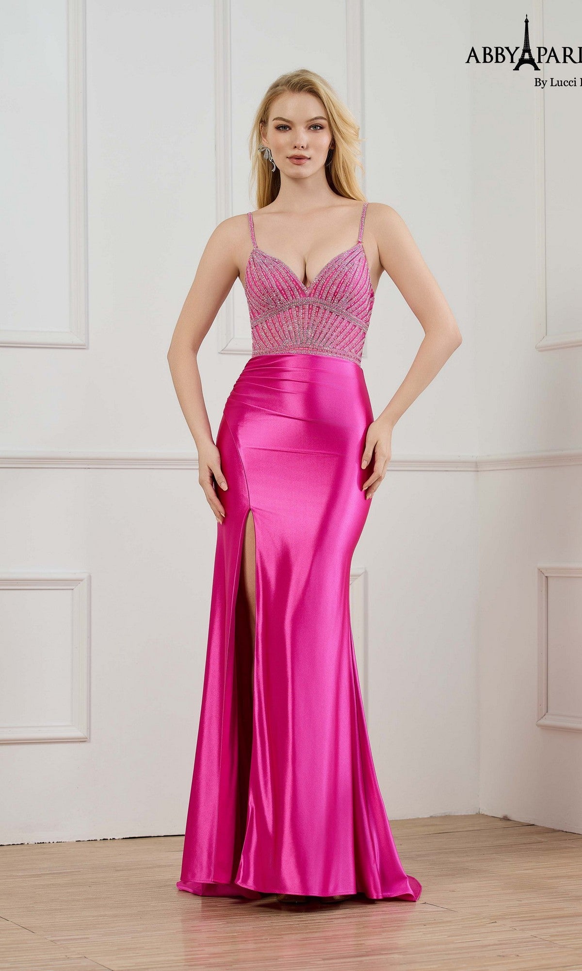 A woman stands against a white wall in the Abby Paris 90364 long prom dress, featuring fuchsia satin, spaghetti straps, a beaded bodice, and a thigh-high slit.