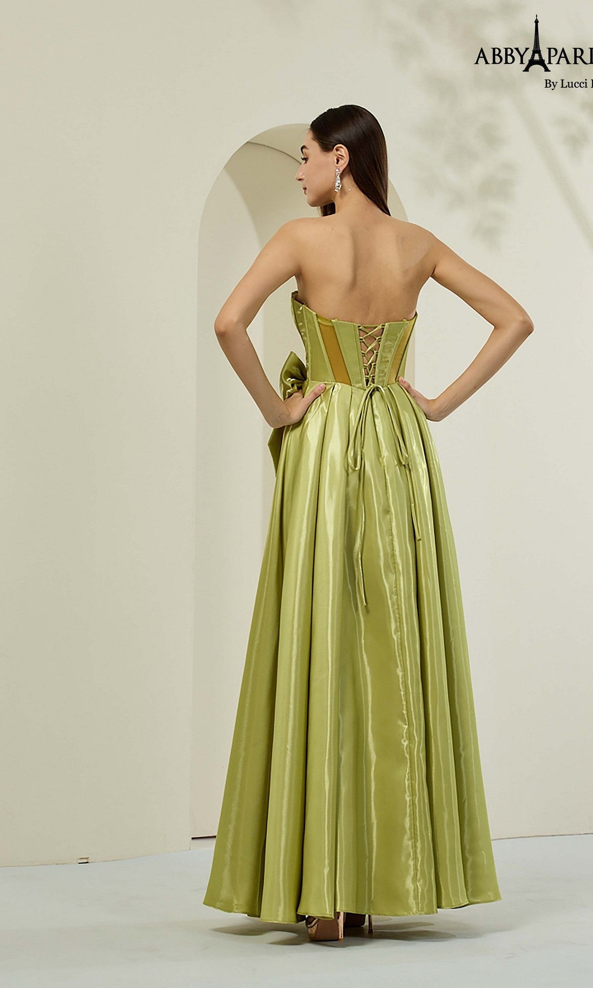A woman faces away in a minimalist setting, wearing the Abby Paris 90384 long prom dress in light green with a strapless corset bodice and lace-up back.