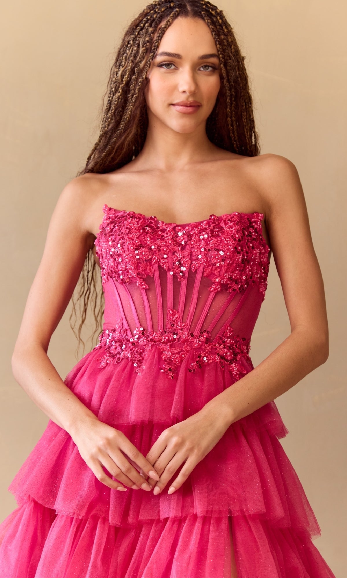 A woman stands against a beige background wearing the Amelia Couture 9053, a strapless long prom ball gown featuring a beaded corset bodice, bright pink floral details, and layered tulle.