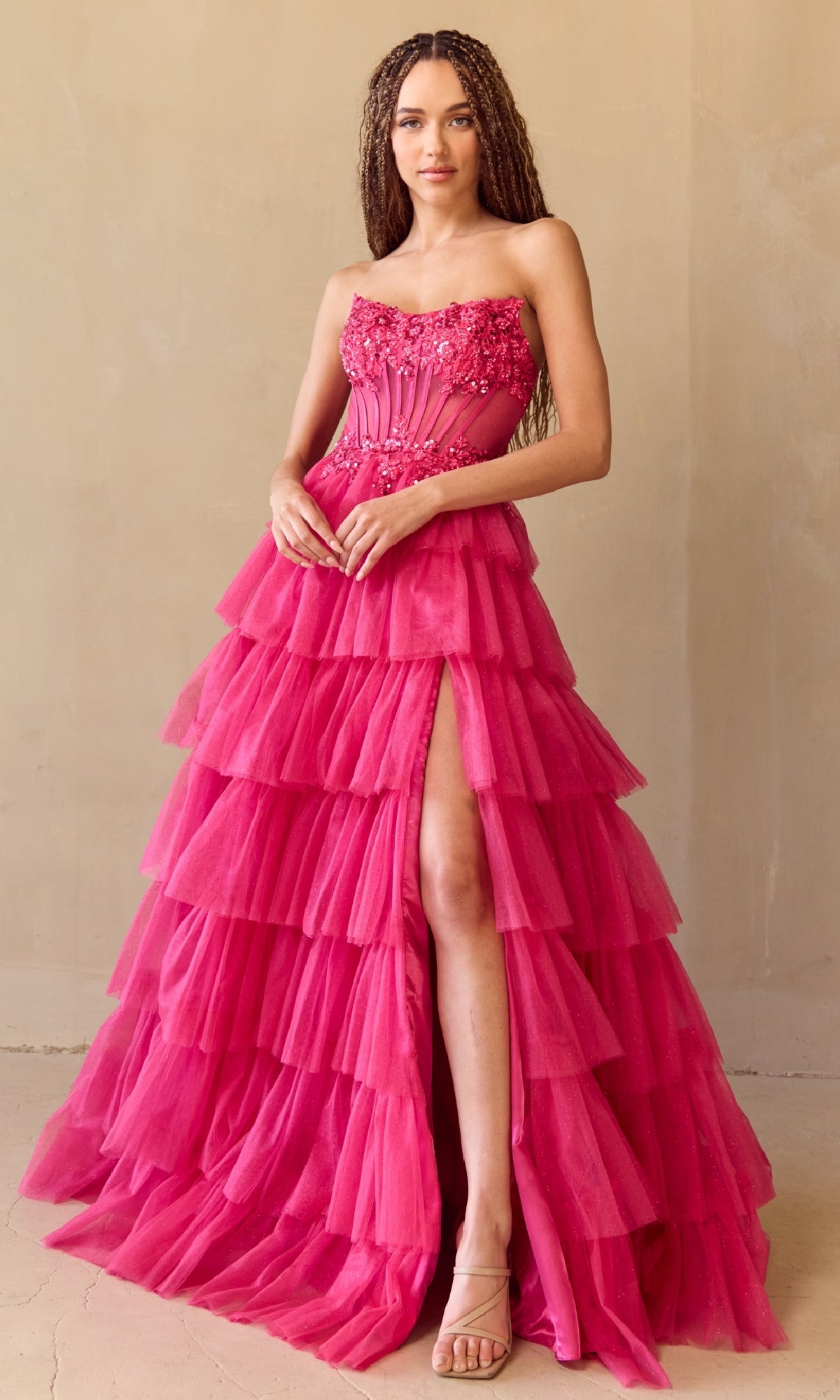 A woman poses against a neutral wall in the Amelia Couture 9053, a strapless long A-line ball gown in bright pink tulle with ruffles, a high slit, and floral beaded embellishments on the corset-style bodice.