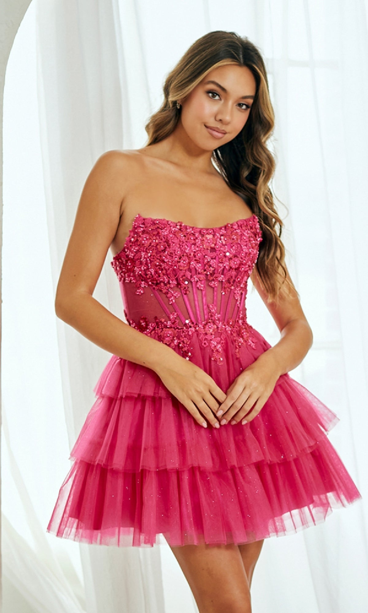 A woman stands indoors wearing the Strapless Ruffle Hoco Dress: Amelia Couture 9066S, a bright pink, strapless dress with floral embellishments and a layered tulle skirt—perfect as a short A-line party dress.