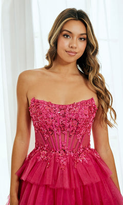 A young woman with long wavy hair stands gracefully before white curtains, wearing the Amelia Couture 9066S Strapless Ruffle Hoco Dress in bright pink, accented with sequins and sheer paneling.