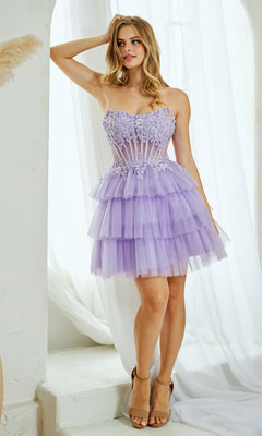 A woman in the Amelia Couture 9066S Strapless Ruffle Hoco Dress, featuring lilac tiers, floral appliqué, and a sheer bodice, stands indoors beside white curtains.