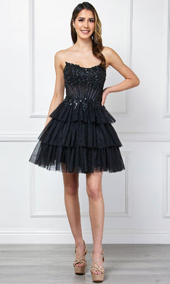 A woman indoors wears the Amelia Couture 9066S Strapless Ruffle Hoco Dress—a black, tiered short A-line party dress with floral embellishments—paired with nude high-heeled sandals.