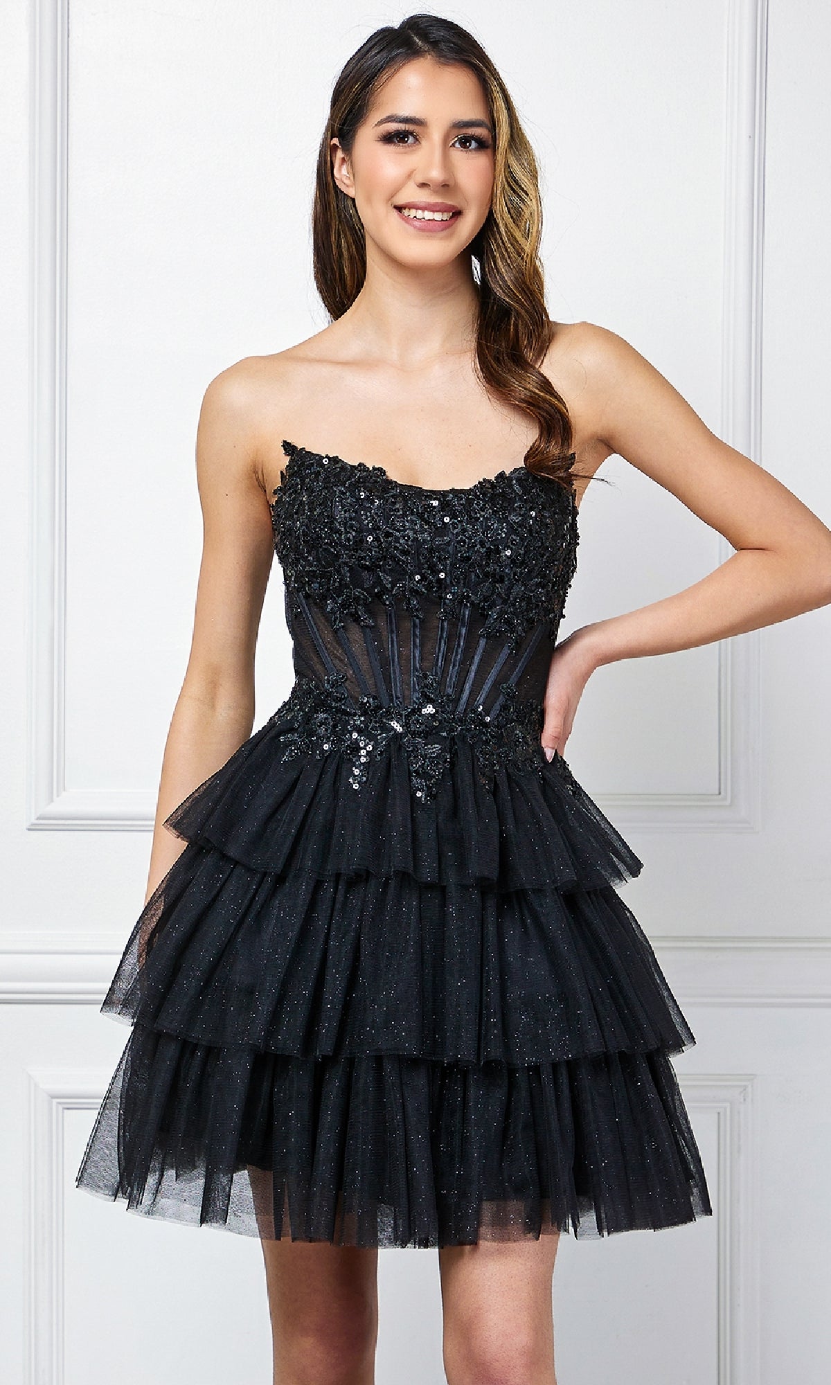 A woman smiles against a white paneled wall, wearing the Amelia Couture 9066S Strapless Ruffle Hoco Dress—a black, tiered mini dress with floral and sequin accents—posing with one hand on her hip.