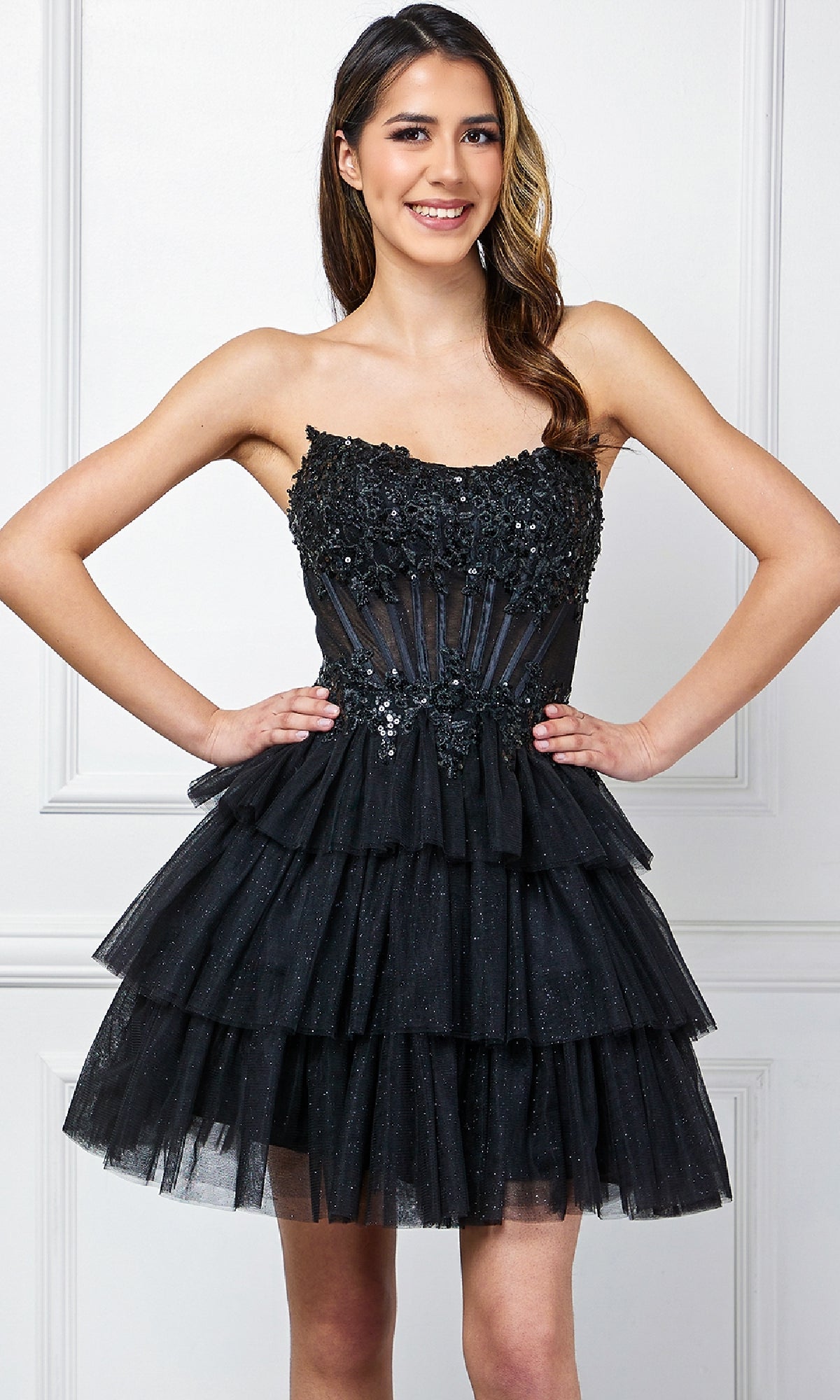 A woman stands smiling against a white paneled wall, wearing the Amelia Couture 9066S Strapless Ruffle Hoco Dress with glitter, floral embellishments, and a layered tulle skirt.