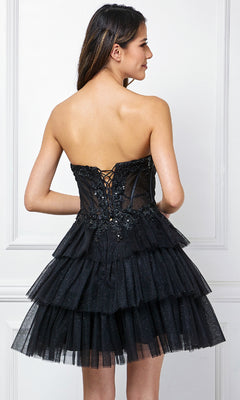 A woman wears the Strapless Ruffle Hoco Dress: Amelia Couture 9066S, featuring a glittery strapless design with a tiered tulle skirt and lace-up corset back, standing by a white paneled wall.