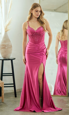 A woman in the Sheer-Waist Long Prom Dress: Amelia Couture 9067—shiny hot pink satin with a beaded corset bodice and thigh-high slit—stands indoors by a large mirror and a black stool topped with a vase.