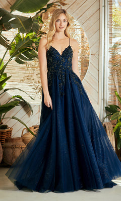 A woman in the Amelia Couture 9079 Lace-Up Long Prom Ball Gown stands indoors by large plants and a wooden wall. The navy blue, floor-length evening dress features spaghetti straps and floral embellishments.