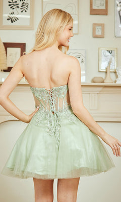 A woman stands facing away in a softly lit room with framed art, wearing the Amelia Couture 9083S strapless short party dress featuring a light green lace-up back and delicate floral details.