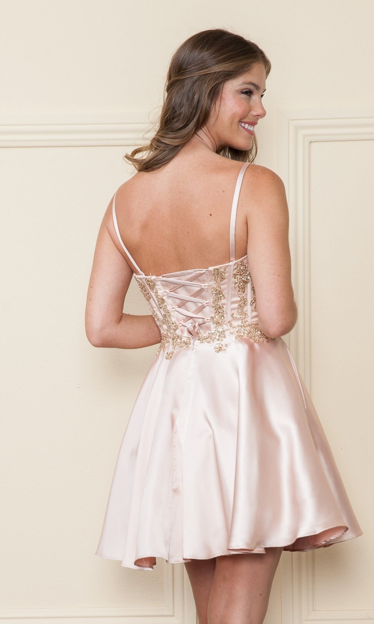 A woman smiles as she faces away from the camera, wearing the Corset-Style Strapless Short Homecoming Dress 9084 in light pink satin with a beaded corset bodice and lace-up back.