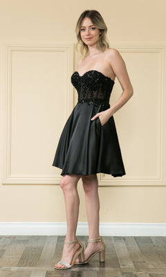 A woman stands indoors wearing the Corset-Style Strapless Short Homecoming Dress 9084, featuring a beaded corset bodice and a flared satin skirt, paired with glittery high-heeled sandals.