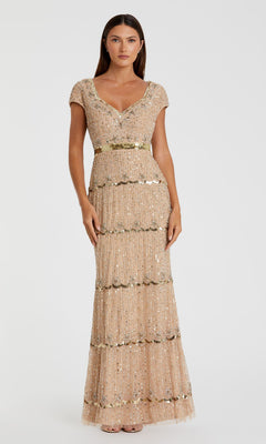 A woman wears the Mac Duggal 9089 Cap-Sleeve Beaded Formal Dress, a floor-length beige gown with gold sequin embellishments and scalloped details, standing against a plain white background.