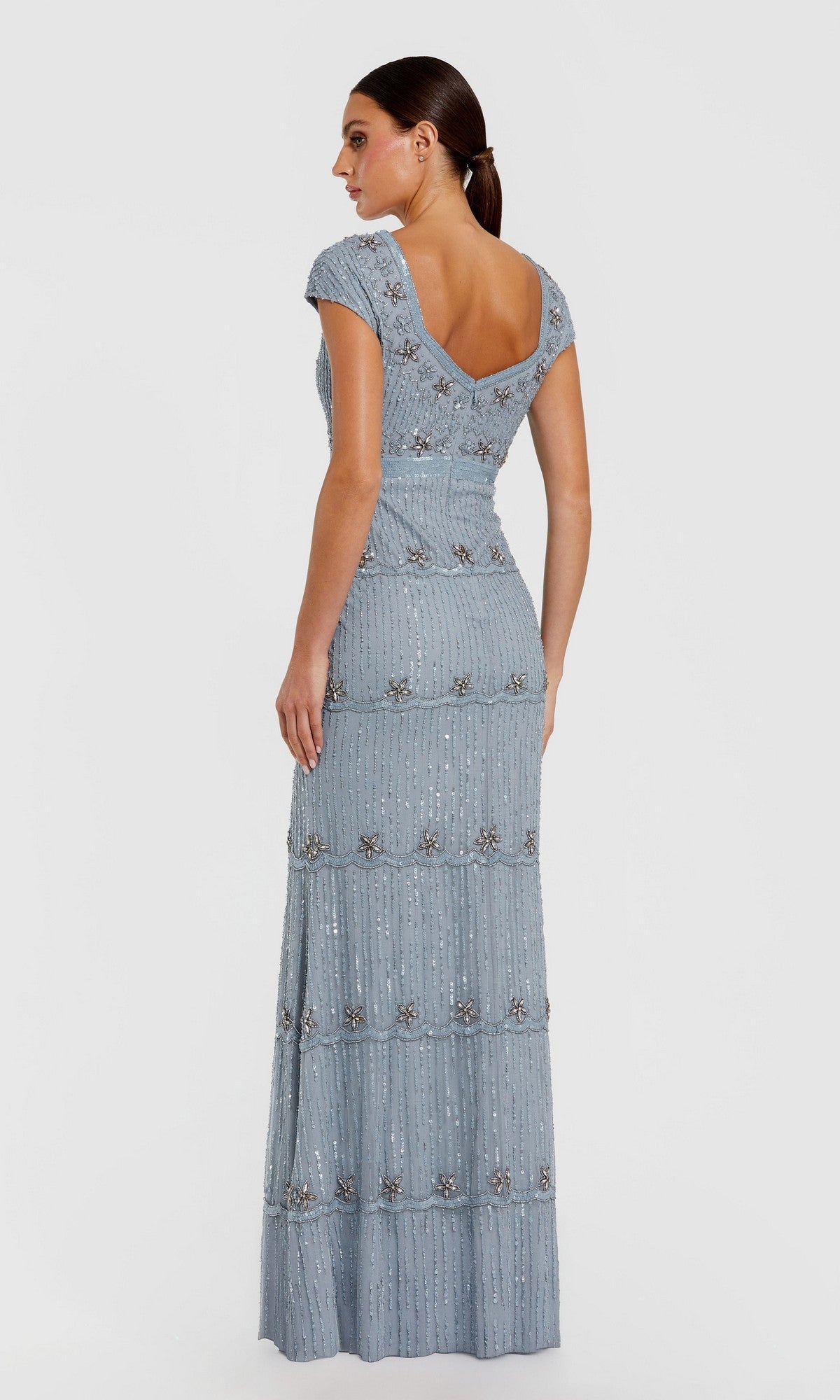 A woman models the Mac Duggal 9089 Cap-Sleeve Beaded Formal Dress in light blue, featuring beaded embellishments and a deep V-back, standing against a plain background.