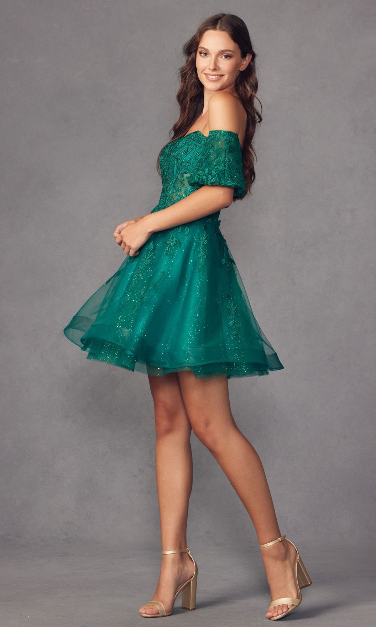 A woman stands sideways, smiling, in the Bell-Sleeve Short Sweetheart Prom Dress 909—an off-the-shoulder green dress with a corset bodice and lace and tulle details—paired with beige block-heeled sandals against a gray background.