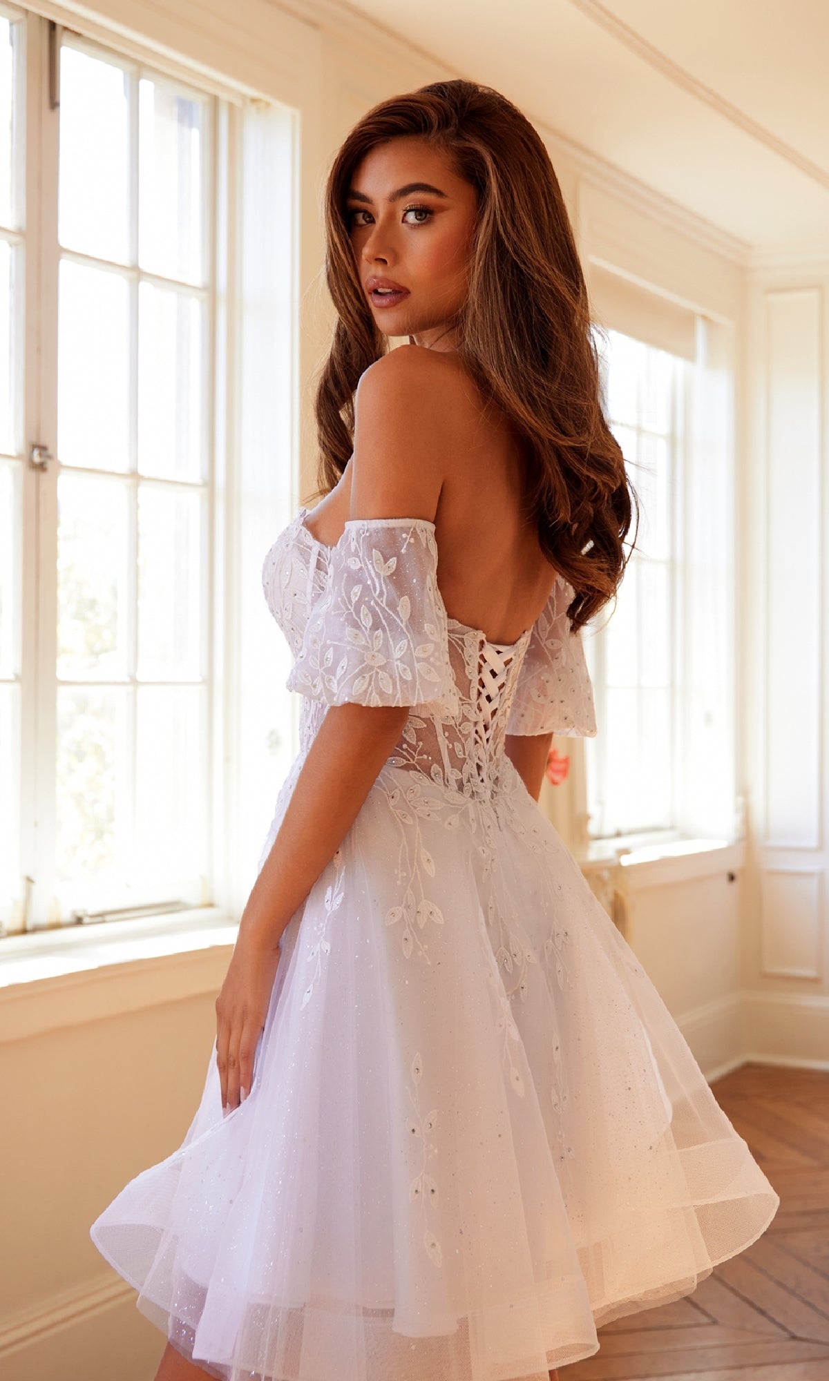 A woman stands in a bright room with large windows, wearing the Bell-Sleeve Short Sweetheart Prom Dress 909—a white, off-the-shoulder dress featuring a corset bodice and floral embroidery.