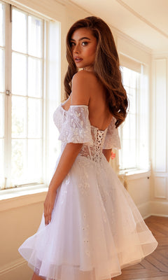 A woman stands in a bright room with large windows, wearing the Bell-Sleeve Short Sweetheart Prom Dress 909—a white, off-the-shoulder dress featuring a corset bodice and floral embroidery.