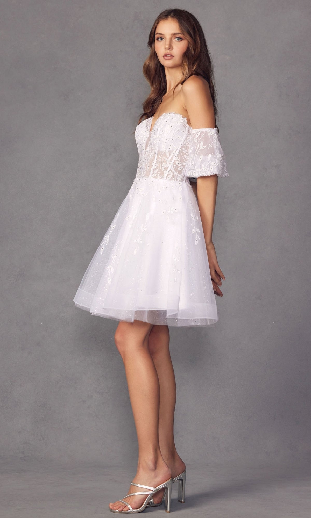 A woman stands in profile wearing the Bell-Sleeve Short Sweetheart Prom Dress 909—white, off-the-shoulder with lace details, sheer sleeves, and floral embroidery—paired with silver heels against a gray background.