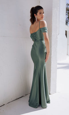 A woman wearing the Classy Long Satin Prom Dress: Eureka 9116 in sage green stands against a white textured wall, elegantly glancing over her shoulder.
