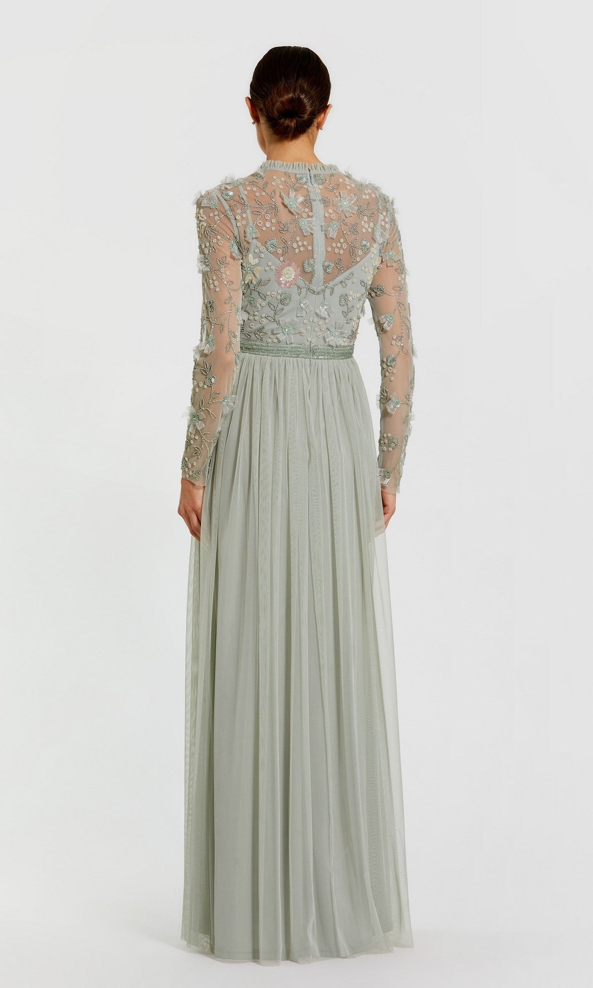 A woman stands facing away, wearing the Mac Duggal 9132 Long Sleeve Vanilla Formal Gown, a floor-length dress featuring sheer embroidered detailing on the bodice and sleeves.