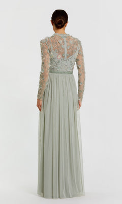 A woman stands facing away, wearing the Mac Duggal 9132 Long Sleeve Vanilla Formal Gown, a floor-length dress featuring sheer embroidered detailing on the bodice and sleeves.