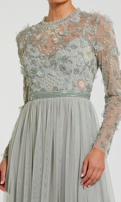 A woman wears the Mac Duggal 9132 Long Sleeve Vanilla Formal Gown featuring sheer long sleeves, a floral embroidered and beaded bodice, and a pleated A-line skirt.