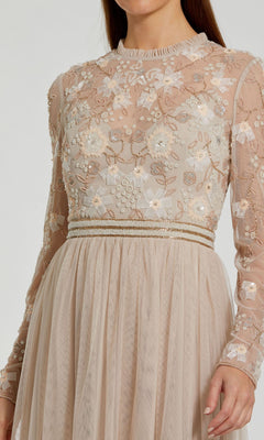 A person wearing the Long Sleeve Vanilla Formal Gown by Mac Duggal 9132—a beige A-line dress featuring floral embroidery and pearl-adorned bodice, with a mesh skirt.