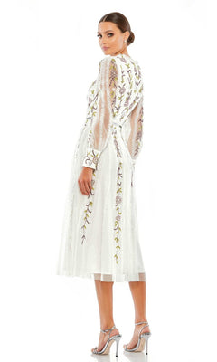 A woman models the Mac Duggal Knee Length Wedding Guest Dress 9133 featuring sheer embroidered sleeves and floral patterns, styled with silver heels—perfect for a semi-formal occasion.