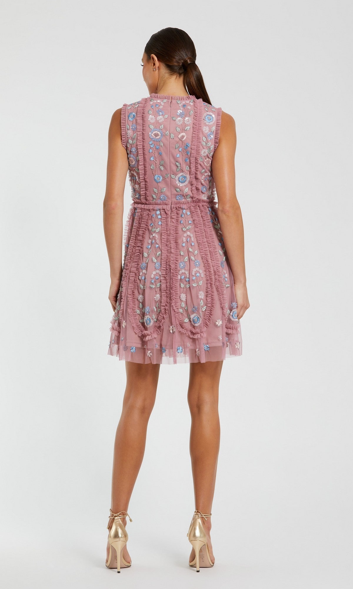 A woman stands facing away, wearing the Mac Duggal 9135 Embroidered Short Party Dress, a sleeveless, knee-length pink floral dress, paired with gold high heels.