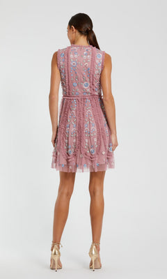A woman stands facing away, wearing the Mac Duggal 9135 Embroidered Short Party Dress, a sleeveless, knee-length pink floral dress, paired with gold high heels.
